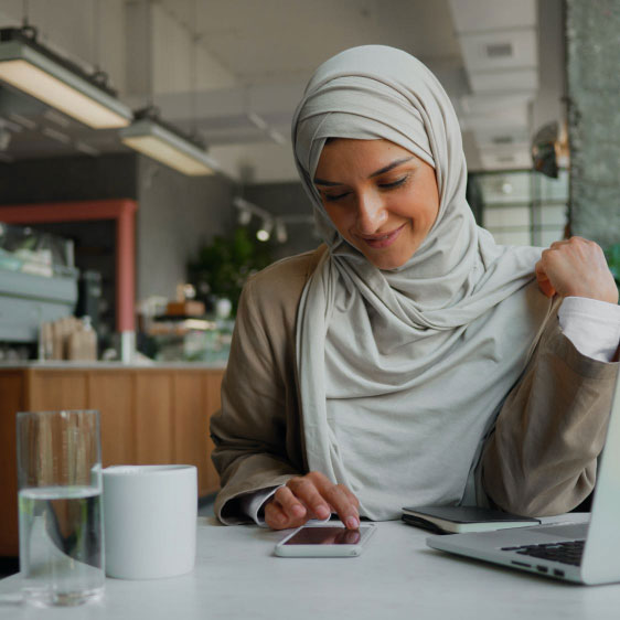 Woman with a hijab sits at a table in a cafe while smiling and glancing down at a phone.
