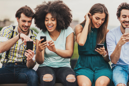null A group of young people sit together, smiling and laughing with each other while looking at their mobile phones.