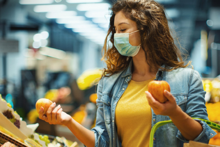 null A young woman wearing a face mask and holding a shopping basket stands in a supermarket comparing two apples in each of her hands.